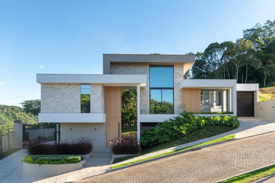 Fachada da Casa JG, com volumes planos em revestimento de pedra, painéis de madeira e grandes vidros, implantada no terreno em declive
